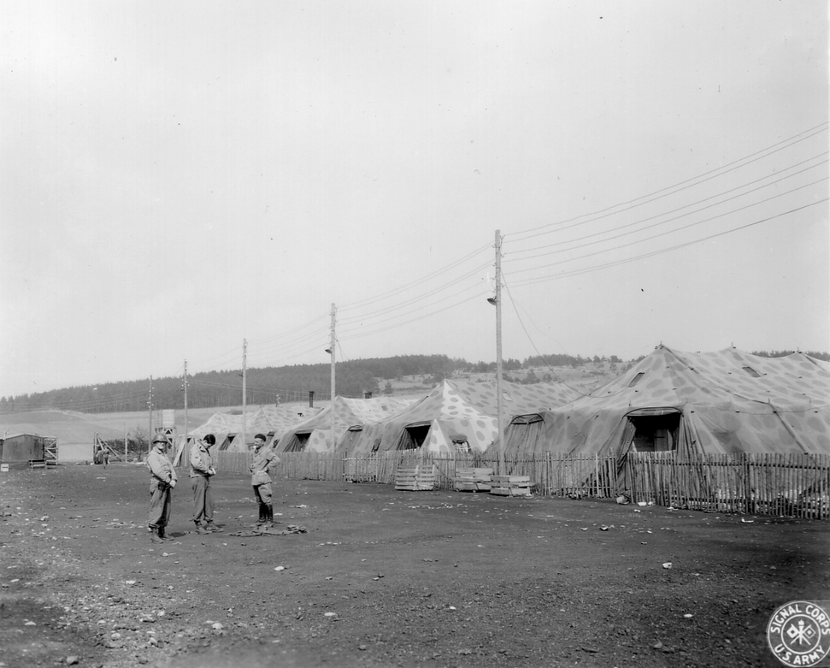 Die getarnten Zelte im Lager Espenfeld, 13. April 1945. Im Vordergrund Soldaten der U.S. Army im Gespräch mit einem Überlebenden. Foto: Arthur W. Stott (U.S. Army Signal Corps)