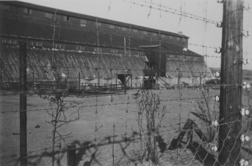Blick durch den Stacheldrahtzaun auf eine der beiden Lagerhallen des ehemaligen Außenlagers in Kleinbodungen, Mai 1945. Das Foto nahm der französische Zivilarbeiter René Tournemire auf.