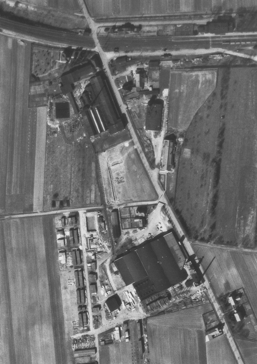 Vergrößerter Ausschnitt der Aufnahme der alliierten Luftaufklärung vom 22. März 1945. Unterhalb der Bahntrasse sind die Gebäude der Mechanischen Weberei AG zu sehen, die als Häftlingsunterkunft dienten, darunter der ehemalige Appellplatz des Lagers und die Rundbogenhallen des Sperrholzwerks Hermann Becher, in denen die Häftlinge arbeiten mussten. (Ausschnitt)