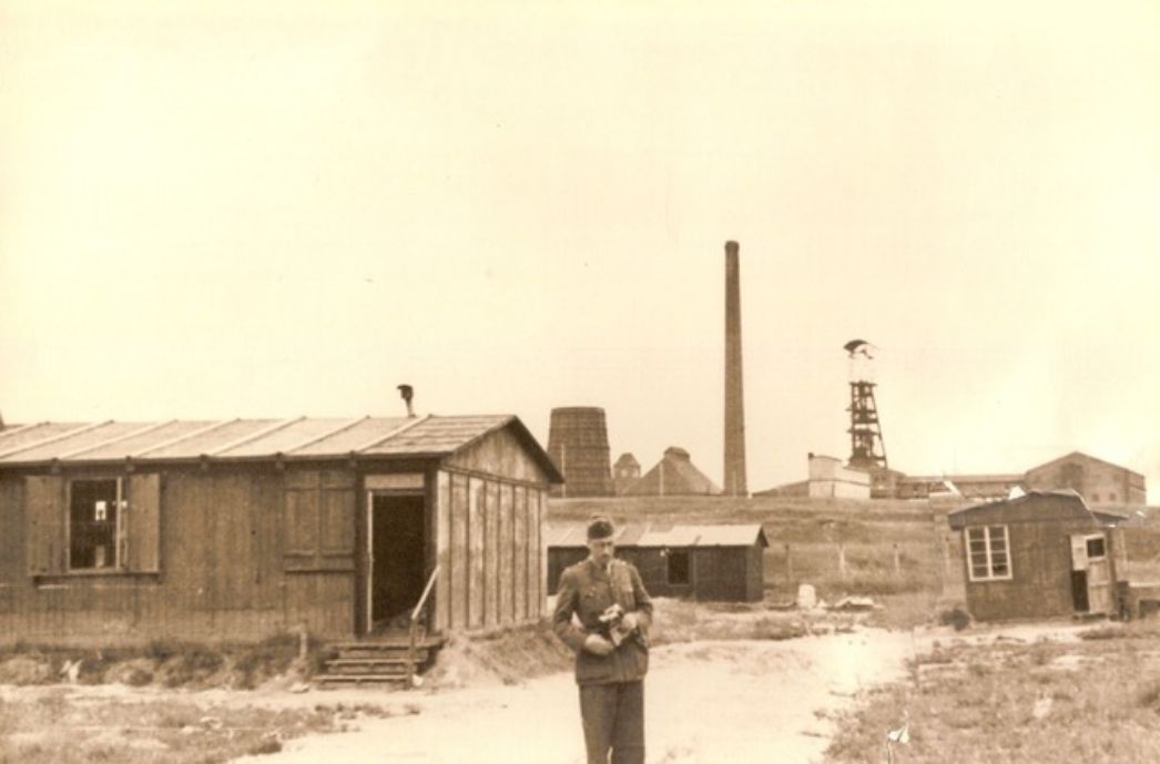 Das ehemalige Barackenlager mit einer der Schachtanlagen im Hintergrund, 1945. Um wen es sich bei dem Uniformierten handelt, ist nicht bekannt.
